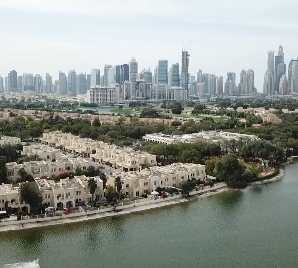 Aerial view of a villa and townhouse community beside a lake, with the Dubai skyline behind